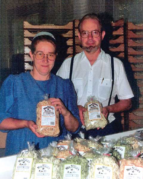Founders Leon and Esther Miller each holding a bag of Mrs. Miller's noodles, standing behind a table of packaged noodles with wooden drying racks behind them