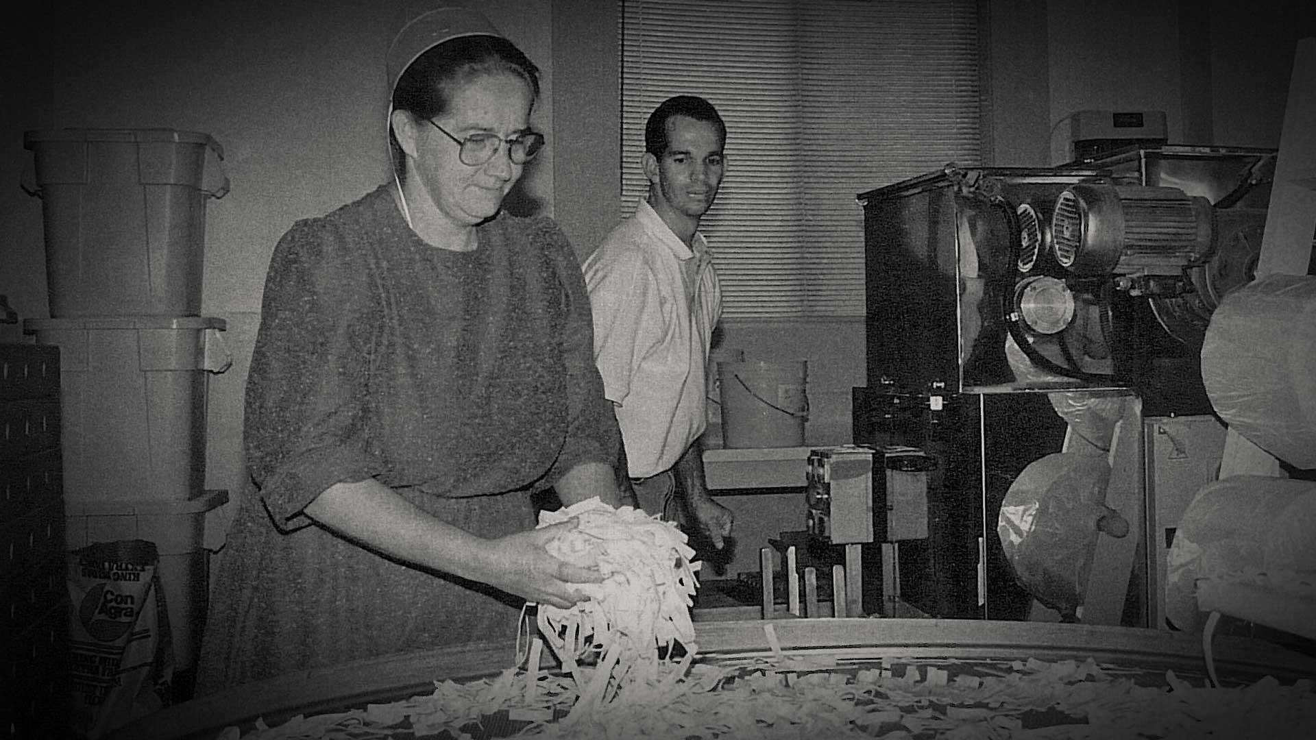 Esther Miller lifting a handful of freshly cut egg noodles from a round drying table, with noodle-making equipment and a helper behind her