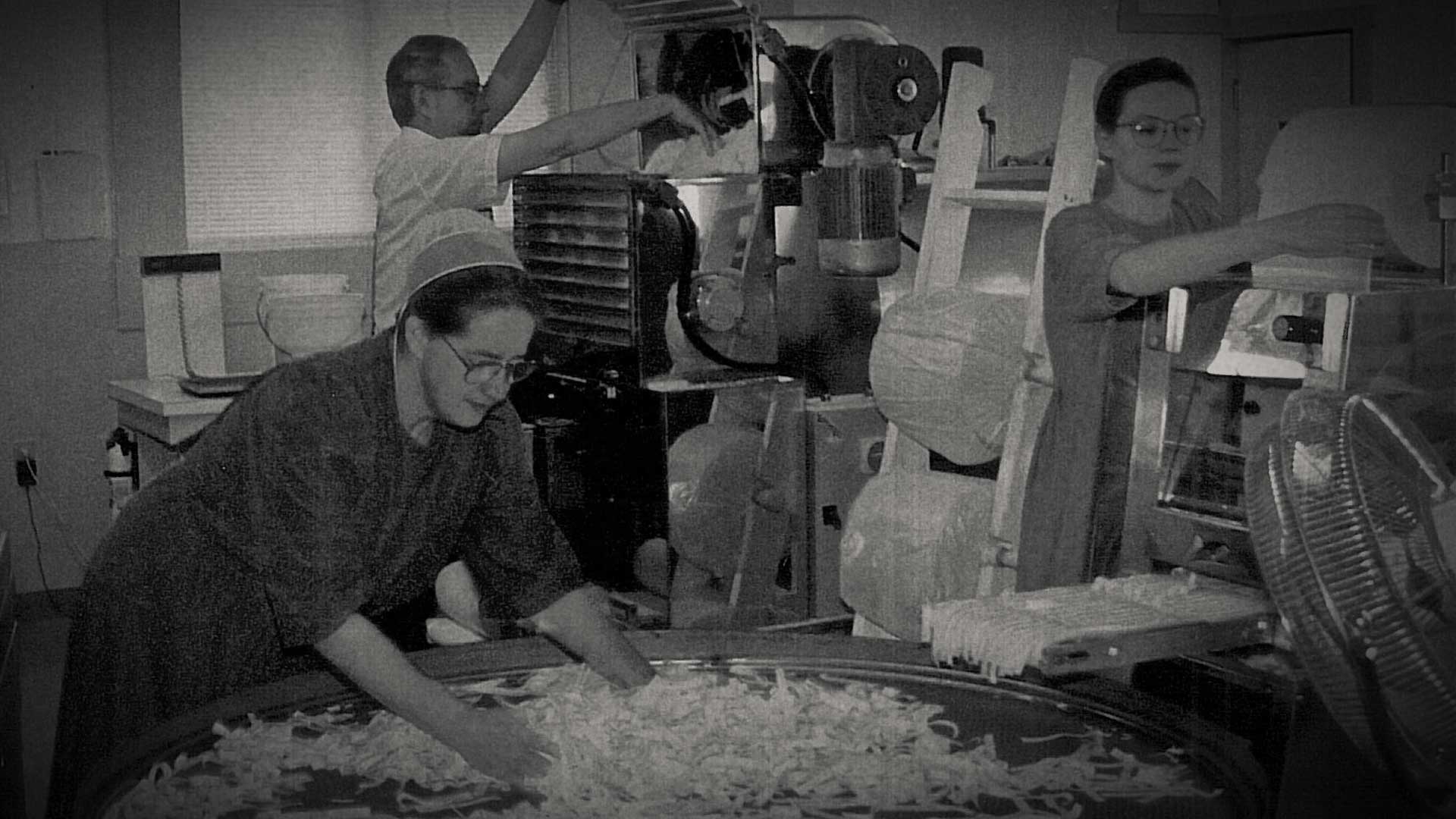 Four members of the Miller family working in the early noodle production room — spreading cut noodles across a round table, tending the rolling equipment, and stacking drying racks